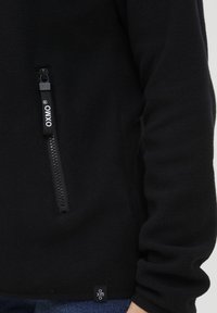 Pull en polaire noir avec une poche zippée à l'avant, texture lisse et une petite étiquette logo près de l'ourlet.