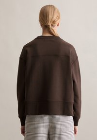 Brązowa bluza z prążkowanym dekoltem i mankietami, o luźnym kroju. Tekstura jest gładka, w połączeniu z spodniami w kratę.