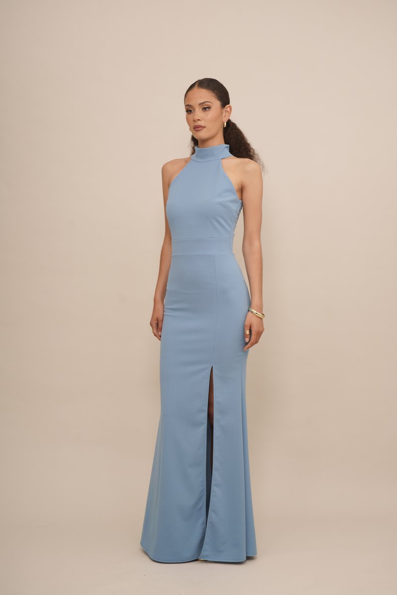 Woman with curly hair wearing a sleeveless blue high-neck gown with a front slit, standing against a plain beige background.
