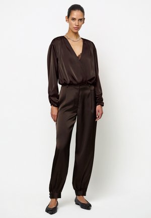 Vrouw draagt een donkerbruine satijnen blouse met lange mouwen en een diepe V-hals en bijpassende satijnen broek met omgeslagen enkels en zwarte platte schoenen.