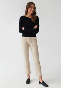 Maglietta nera a maniche lunghe e pantaloni beige a gamba dritta con dettagli di cucitura visibili. Scarpe nere slip-on completano l'outfit.