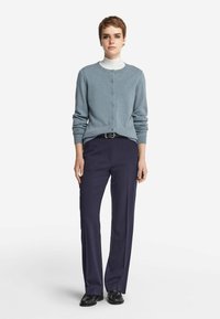 Niebieski kardigan z guzikami, zestawiony z ciemnoniebieskimi spodniami o prostym kroju i czarnymi butami. Stylizacja zawiera warstwowy biały golf noszony pod spodem.