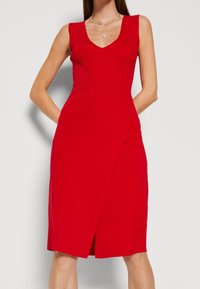 Robe rouge sans manches avec un décolleté en V, un corsage côtelé, une taille cintrée et une fente sur le côté. Confectionnée dans un tissu lisse avec une silhouette élégante.