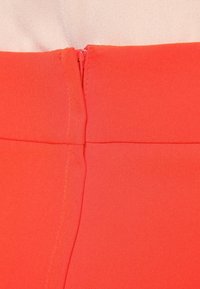 Červený látkový materiál s hladkou texturou a vysokým leskem. Má skrytý zip na zadní straně a těsně přiléhající střih v pase.