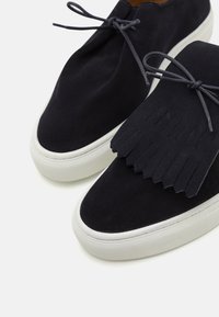 Schwarze Wildleder-Sneaker mit fransigen Akzenten, weißer Gummisohle und passenden Schnürsenkeln. Das Design verfügt über eine abgerundete Zehenpartie und eine glatte Textur.