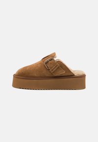 Brown suede slip-on clogs feature a cushioned lining, a wide strap with a buckle accent, and a thick platform sole with a zigzag edge.