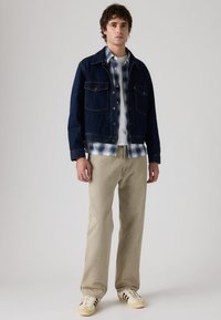 Μπουφάν τζιν σε σκούρο μπλε χρώμα με δύο τσέπες στο στήθος, φορεμένο πάνω από πουκάμισο καρό σε μπλε και λευκό χρώμα. Μπεζ παντελόνι και λευκά αθλητικά παπούτσια με καφέ ρίγες.