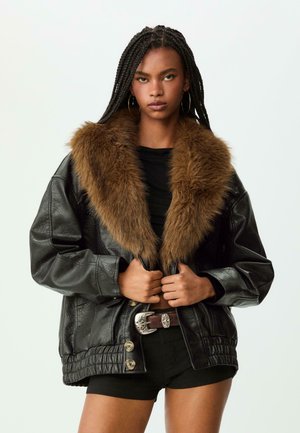 Schwarze Lederjacke mit einem braunen Kunstpelzkragen, gestepptem Saum und großen Knöpfen. Das Modell trägt ein schwarzes Oberteil und Shorts mit einem dekorativen Gürtel.