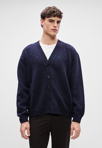 Sötétkék kötött kardigán, V nyakú dizájnnal, bordázott mandzsettákkal és szegéllyel, öt fekete gombbal az elején. Puha tapintású és laza szabású.
