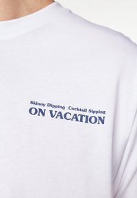 Biała koszulka z granatowym napisem "Nocne kąpiele, koktajle na wakacjach" na górnej części klatki piersiowej.