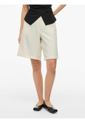 Nő krémszínű, térdig érő rövidnadrágban, fekete felsővel és fekete lapos cipővel, fehér háttér előtt állva.