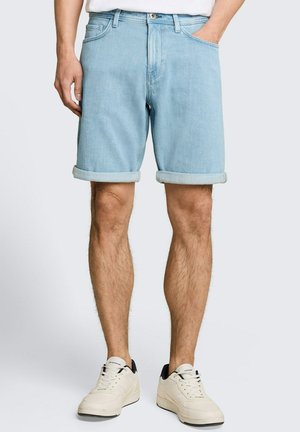 Man draagt lichtblauwe opgerolde denim shorts, witte sneakers met zwarte details en een wit overhemd, staand met ontspannen handen.