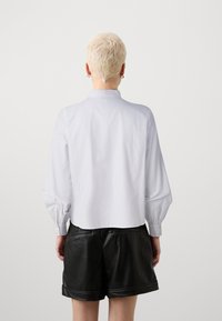 Camicia bianca a maniche lunghe con colletto alto e polsi abbottonati, abbinata a pantaloni corti in pelle nera con orlo arrotolato e tasche posteriori.