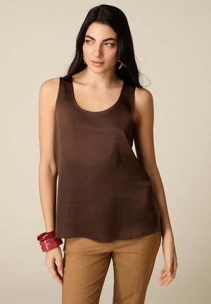 Donna con lunghi capelli neri che indossa una canotta marrone scuro, pantaloni beige, orecchini d'oro e braccialetti rossi al polso sinistro.