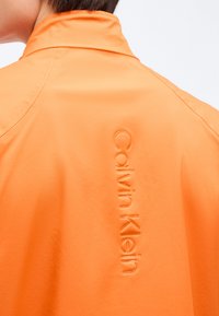 Ώμος και άνω μέρος της πλάτης ενός ατόμου που φοράει πορτοκαλί μπουφάν με ανάγλυφο κάθετο κείμενο "Calvin Klein" στην πλάτη.