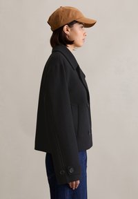 Manteau peacoat en mélange de laine noir avec un col en pointe, des poches latérales et des poignets boutonnés, associé à une casquette de baseball beige et un jean en denim.