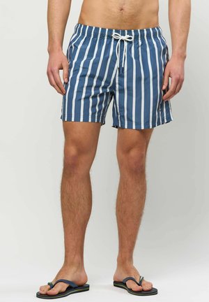 Mann trägt blaue und weiße vertikal gestreifte Badeshorts mit weißem Kordelzug, steht barfuß in marineblauen Flip-Flops vor einem einfarbigen Hintergrund.