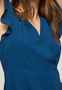 Modrá texturovaná blůza s výstřihem do V a volnými krátkými rukávy. Tkanina má jemný povrch, který zvýrazňuje její vrstvený design.