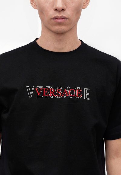 Άτομο που φοράει μαύρο μπλουζάκι με τη λέξη Versace κεντημένη δύο φορές σε επικαλυπτόμενα λευκά και κόκκινα γράμματα στο στήθος.