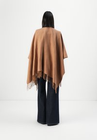 Brun vævet poncho med et struktureret mønster og frynset kant, båret over mørke flagrende jeans. Vist mod en helt hvid baggrund.