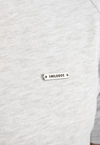 Lys grå stof t-shirt med et lille sølvmetal mærke med teksten "SMILODOX," fastgjort med to synlige nitter.