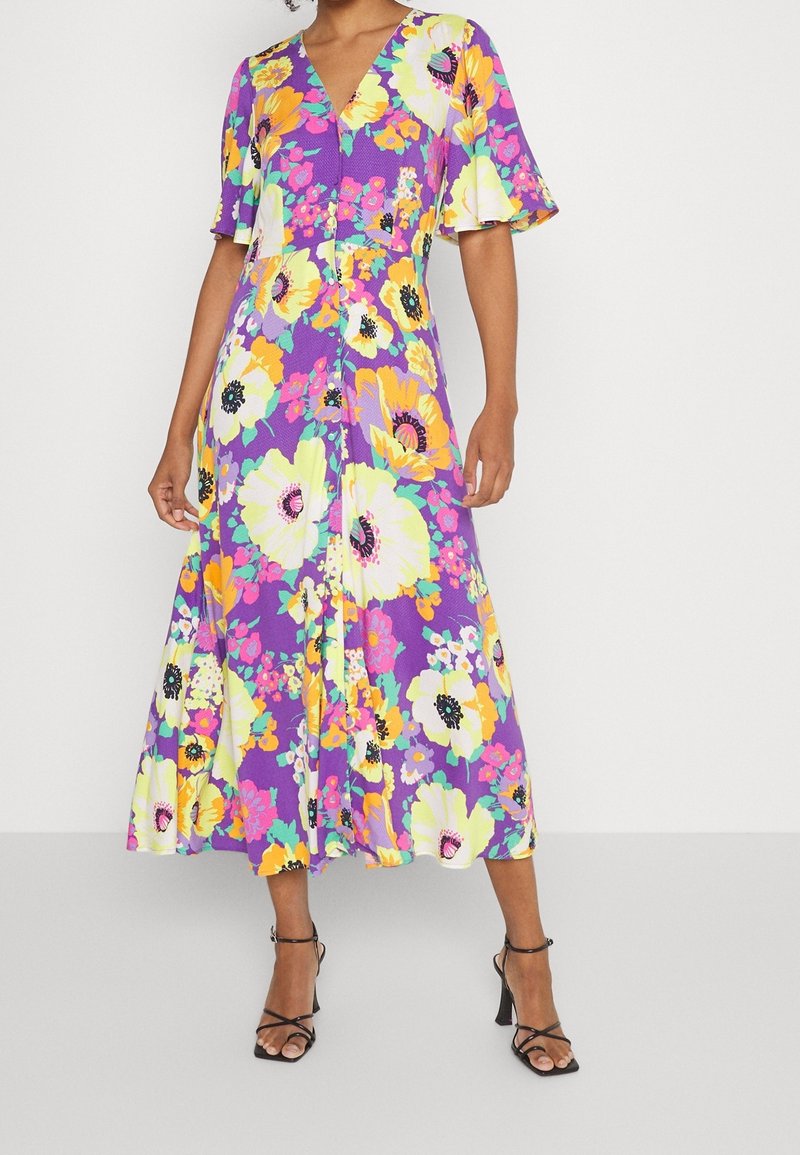 Abito maxi floreale con sfondo viola e fiori gialli, rosa e verdi. Collo a V, maniche corte a fluttuazione e silhouette fluida.
