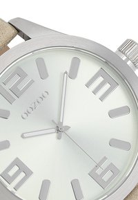 Montre en argent avec un cadran vert clair réfléchissant, dotée de grands marqueurs horaires et d'aiguilles lisses et minces. Bracelet beige à finition texturée.
