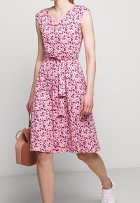 Růžová květinová šaty s výstřihem do V, krátkými rukávy a páskem v pase. Obsahuje vzor listů a rozšířenou sukni. Nosené s bílými teniskami a růžovou taškou.