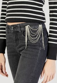Vaqueros de mezclilla negra con un diseño ajustado, que cuentan con cadenas de rhinestones en cascada en el bolsillo y una textura sutil. Blusa a rayas en blanco y negro.
