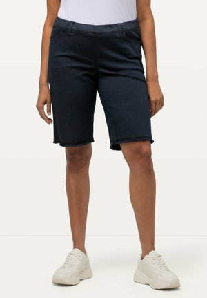 Bermudas en denim bleu foncé avec une taille élastique, présentant un ourlet effiloché et deux poches avant. Portées avec des baskets blanches.