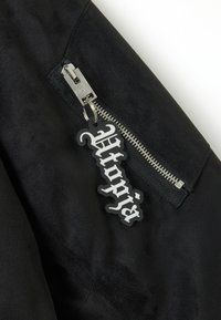 Giacca in suede nera con zip argentata e un'etichetta di plastica con la scritta "Utopia" in carattere stilizzato bianco attaccata alla zip.