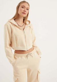 Felpa beige tagliata con zip frontale, cappuccio e due tasche frontali. Abbinata a pantaloni beige coordinati con tasche laterali cargo.