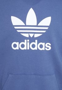 Blå bomullströja med en stor vit Adidas-logotyp, tre ränder och en treklövergrafik på framsidan, inklusive en känguruficka.