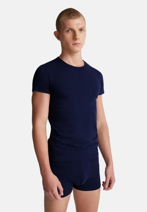 Młody mężczyzna ubrany w dopasowany granatowy t-shirt z krótkim rękawem oraz pasujące granatowe bokserki na jednolitym tle.