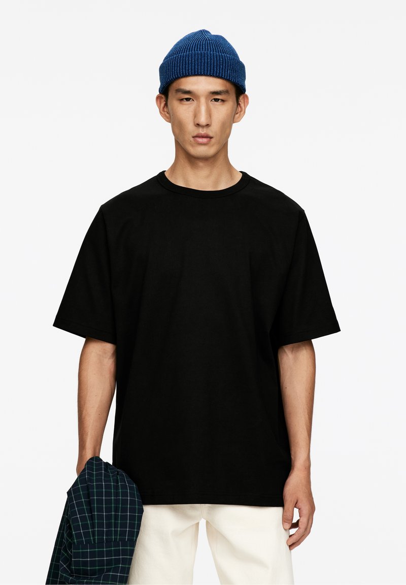 En ung man som bär en blå stickad mössa och en svart oversized t-shirt, som håller en blå och grön rutig skjorta, mot en vit bakgrund.