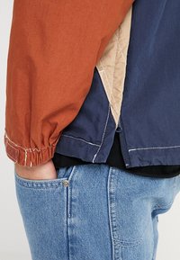 Kleurvlakkenjas met delen in roestoranje, marineblauw en beige. Voorzien van elastische manchetten, contrasterende stiksels en een half-ritssluiting aan de zoom.