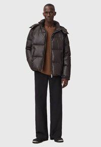 Mand iført en sort pufferjakke over en brun striktrøje, sorte vide bukser og sorte slip-on sko, som står imod en grå baggrund.