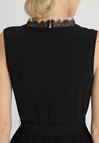 Abito nero senza maniche con colletto in pizzo, chiusura zip sulla schiena e vita aderente con una gonna fluente. Tessuto dalla texture liscia.