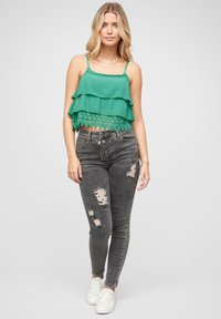 Zeleni slojeviti crop top s čipkastim rubom, uparen s iznošenim crnim skinny trapericama i bijelim tenisicama. Glatka tkanina, prilagođeni kroj i teksturirani detalji.