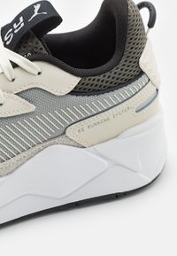 Lichtbeige sneaker met bovenwerk van mesh en stof, geaccentueerd met textuur in grijs, witte tussenzool en "RS Running System" detail aan de zijkant.