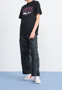 Černé tričko Nike s růžovým logem, doplněné širokými černými kalhotami s květinovým vzorem. Na nohou jsou bílé tenisky s červenými detaily.