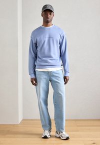 Sudadera azul claro con puños y dobladillo acanalados, superpuesta a una camisa blanca, combinada con jeans azul claro y zapatillas grises. Gorra con logo.