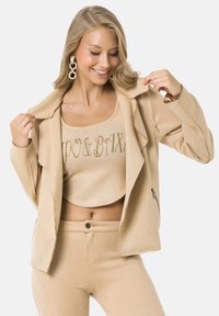 Veste beige à la texture douce avec un design drapé, assortie d'un crop top à ornements dorés et de pantalons ajustés.