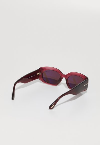 Óculos de sol com tonalidade vermelha, de design elegante e arredondado. Lentes roxas escuras e acabamentos pretos brilhantes nas hastes. Detalhe em metal de lado.