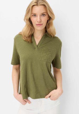 Femme aux cheveux blonds portant une chemise ample manches courtes vert olive avec une poche poitrine décorative, associée à un pantalon blanc.