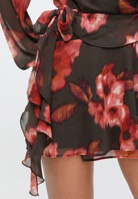 Falda cruzada floral con fondo marrón oscuro, patrones de flores en rosa y rojo, dobladillo con volantes y detalle de lazo en la cintura.