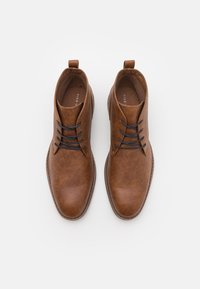 Botas de cuero marrón hasta el tobillo con puntas redondeadas, que presentan cordones oscuros, una pestaña de tirón en forma de bucle y una superficie lisa y texturizada.