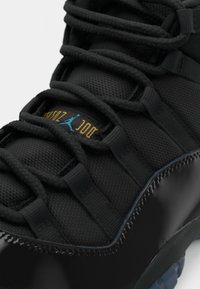 Fekete sportcipő textúrált felsőrésszel, nylon fűzőkkel és fényes fekete patent bőrből készült orral. Az oldalán kék és arany logó található.