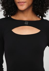 Schwarzes Langarmoberteil mit einem einzigartigen Ausschnittdesign am Hals. Verfügt über ein kleines Logo-Accent in der Nähe des Kragens. Glatte Stoffstruktur.