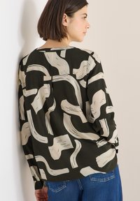 Tmavě zelená blůza s jemným abstraktním vzorem tahů štětcem; dlouhé rukávy, volný střih, zapínání na knoflíky na manžetách, hladká látka. Nošeno s modrými džínami.